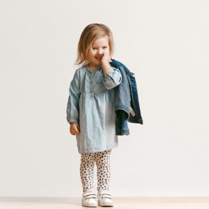 Full length portrait of cute little kid girl in stylish jeans clothes looking at camera and smiling, standing against white studio wall. Kids fashion concept