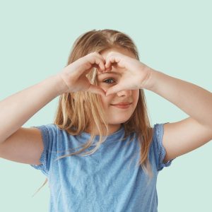 Close up of beautiful blonde girl making heart with hands, looking through it in camera, posing for photo with smile and happy expression